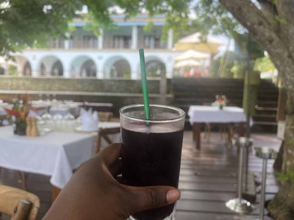 a dark skin woman's hand holding a glass of maroon colored bissap (hibiscus drink)