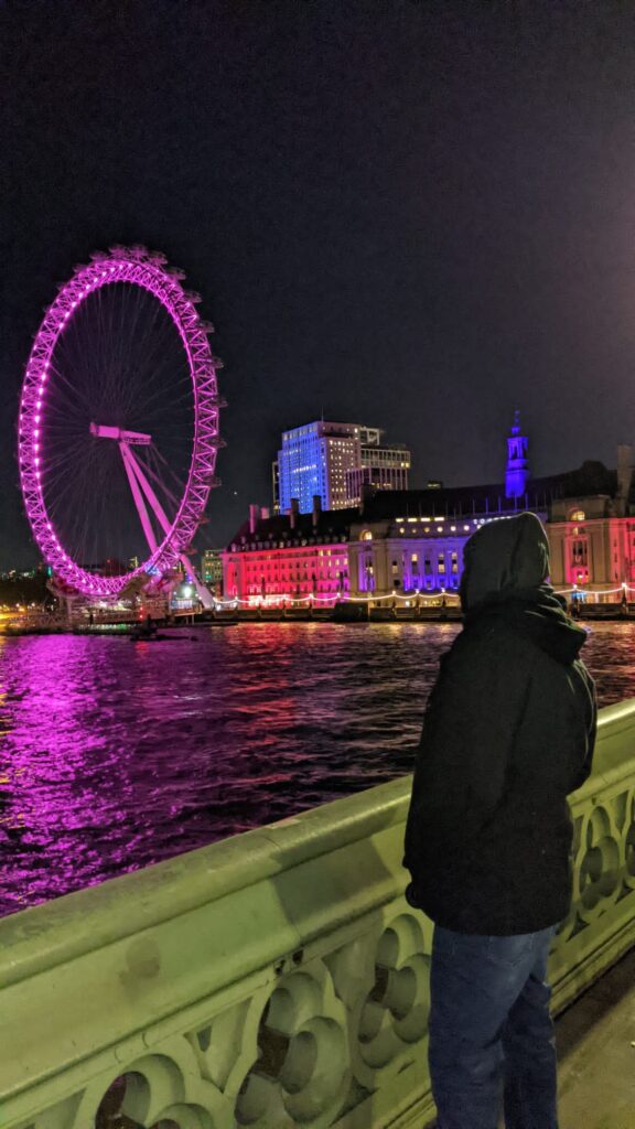 a man with a black jacket and grey hoodie looking at the London Eye Ferris wheel at night. The wheel is lit a bright pink