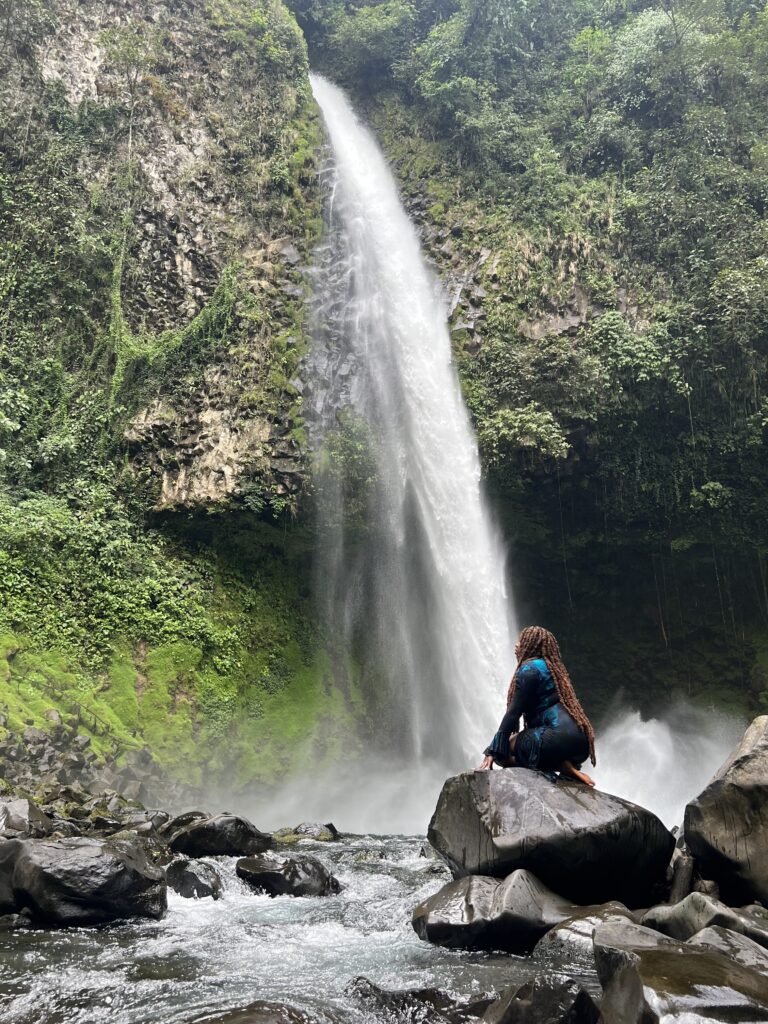 a Black woman kneeling in front of La Fortuna Waterfall