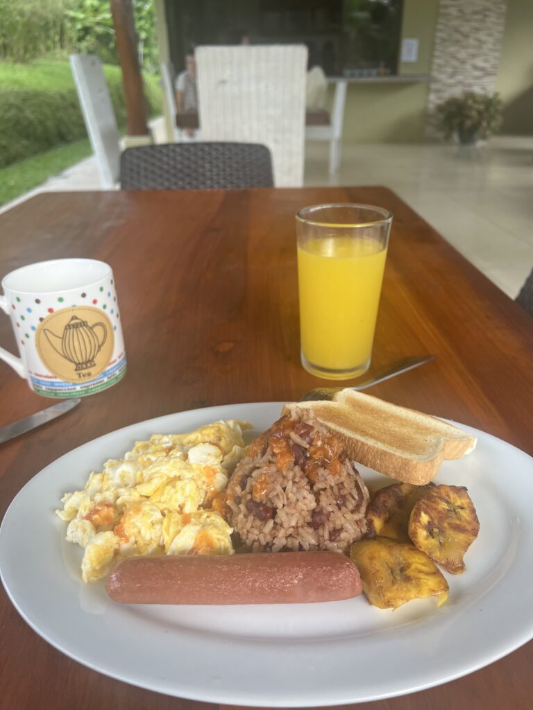 a plate of typical Costa Rican breakfast including a hot dog/sausage, eggs, rice and beans, sweet plantain, bread, orange juice, and coffee