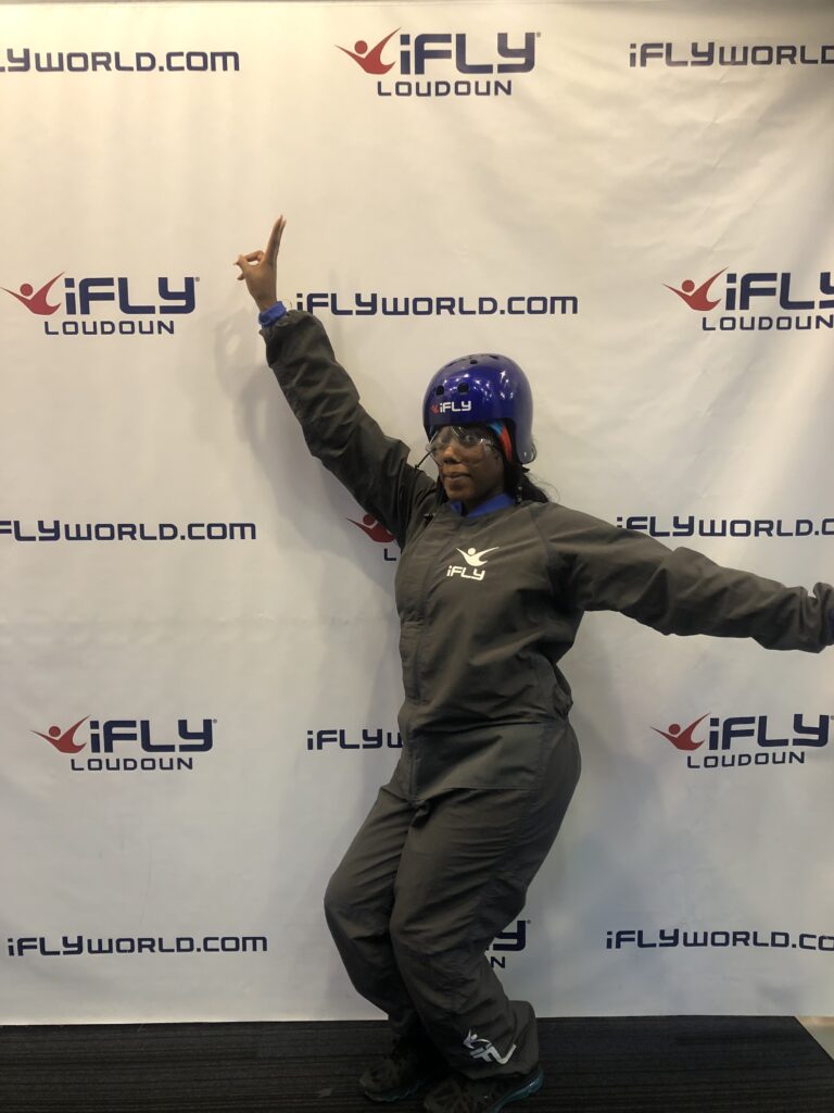a Black woman in an indoor skydiving suit and helmet throwing up the peace sign