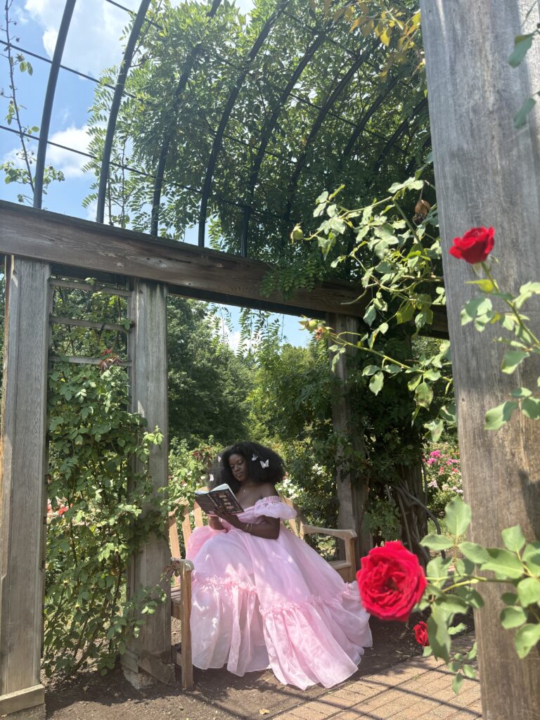 A dark skinned Black woman with large afro and butterflies in her hair reading a book on a bench in Bon Air Rose Garden memorial under a garden pavilion wearing a Pink Selkie dress