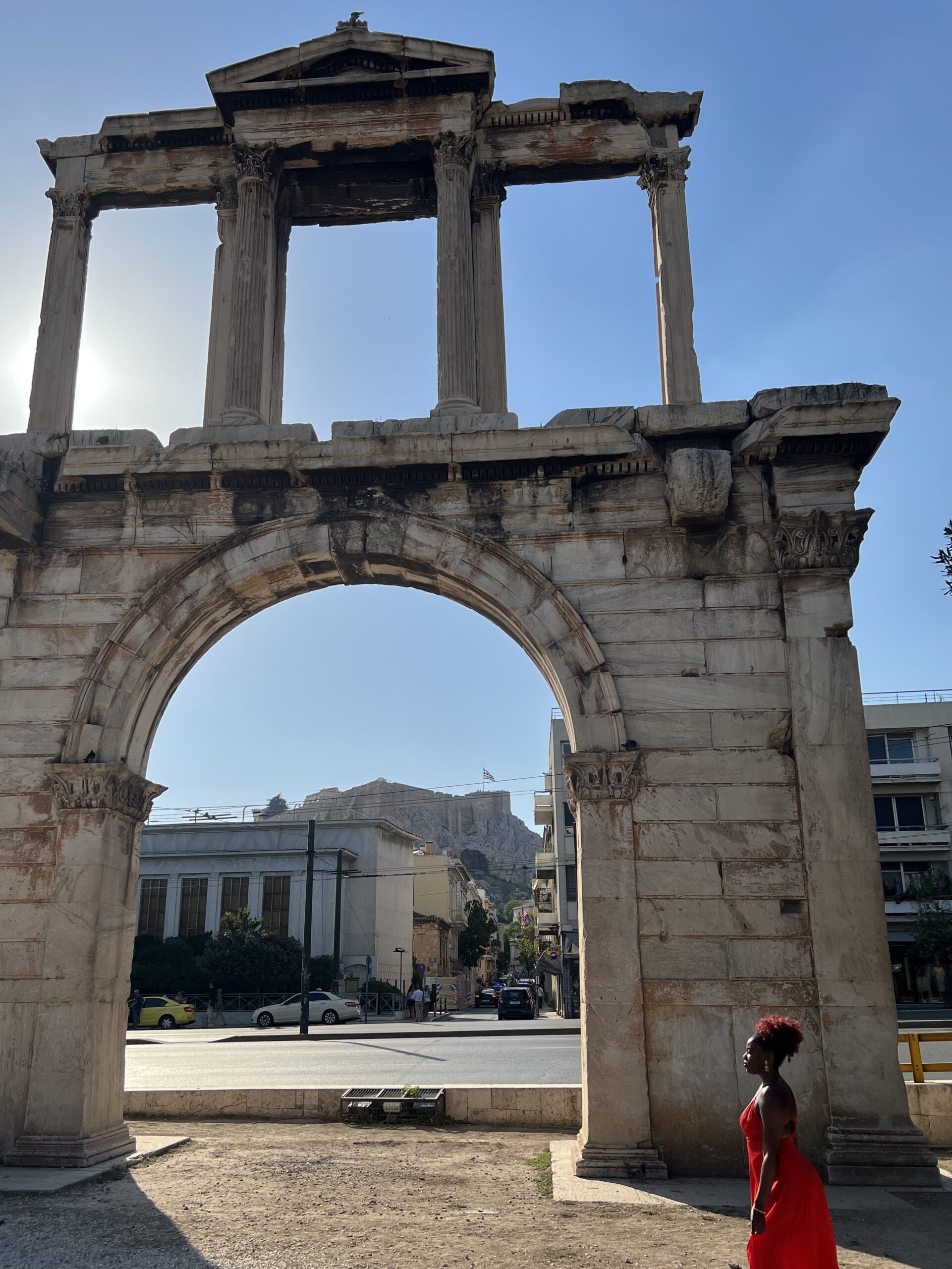 Black woman in red dress and red afro puff standing in front of the ancient Arch of Hadrian in Athens