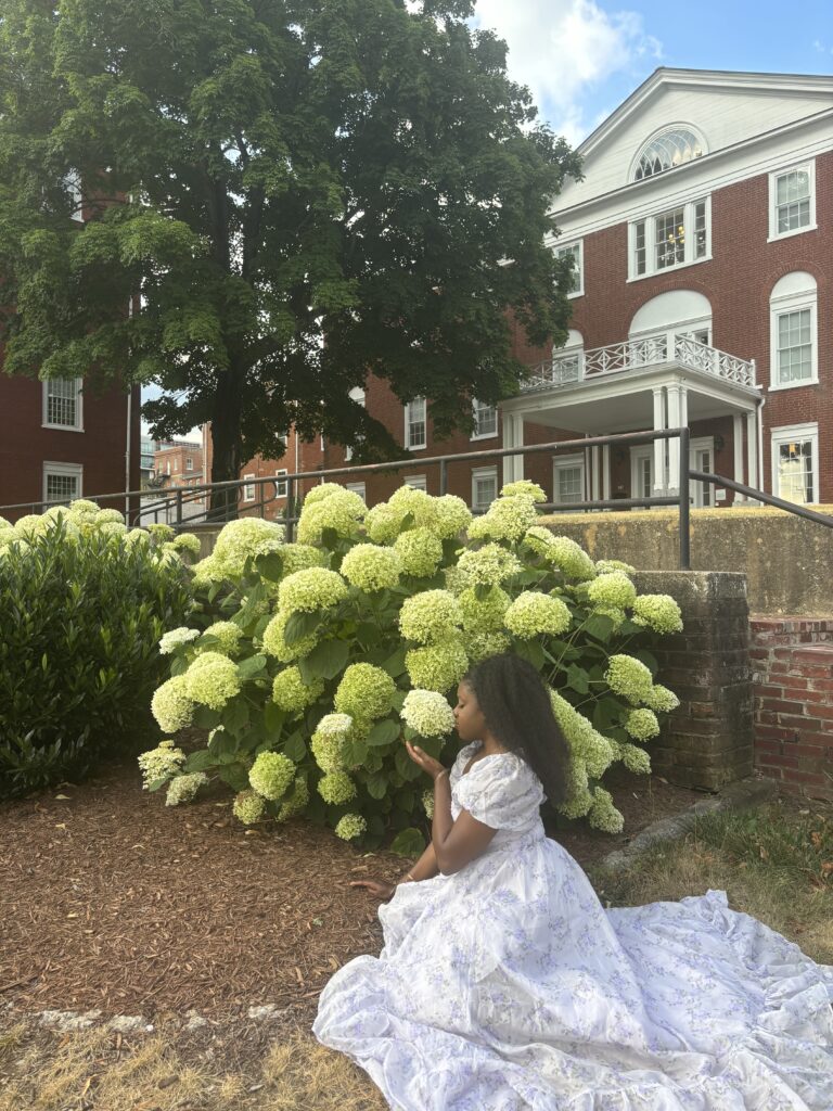 Dark skinned woman with  kinky curly hair sitting in front of a tree with white poofy flowers, while smelling them at Blackburn Inn 