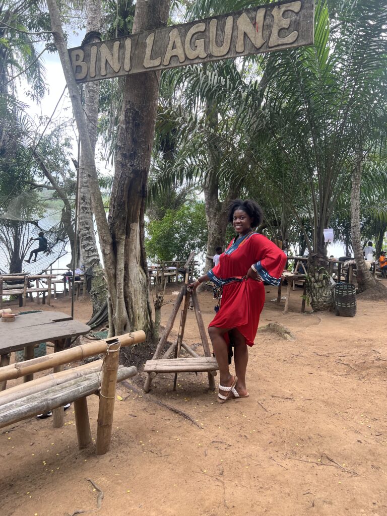 Black woman in red African dress standing in front a sign that says Bini Lagune