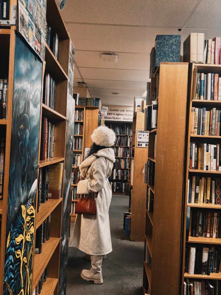 a Black woman in a white coat, white furry hat, white boots, and red purse standing between bookshelves at Second Story Bookstore in Washington DC