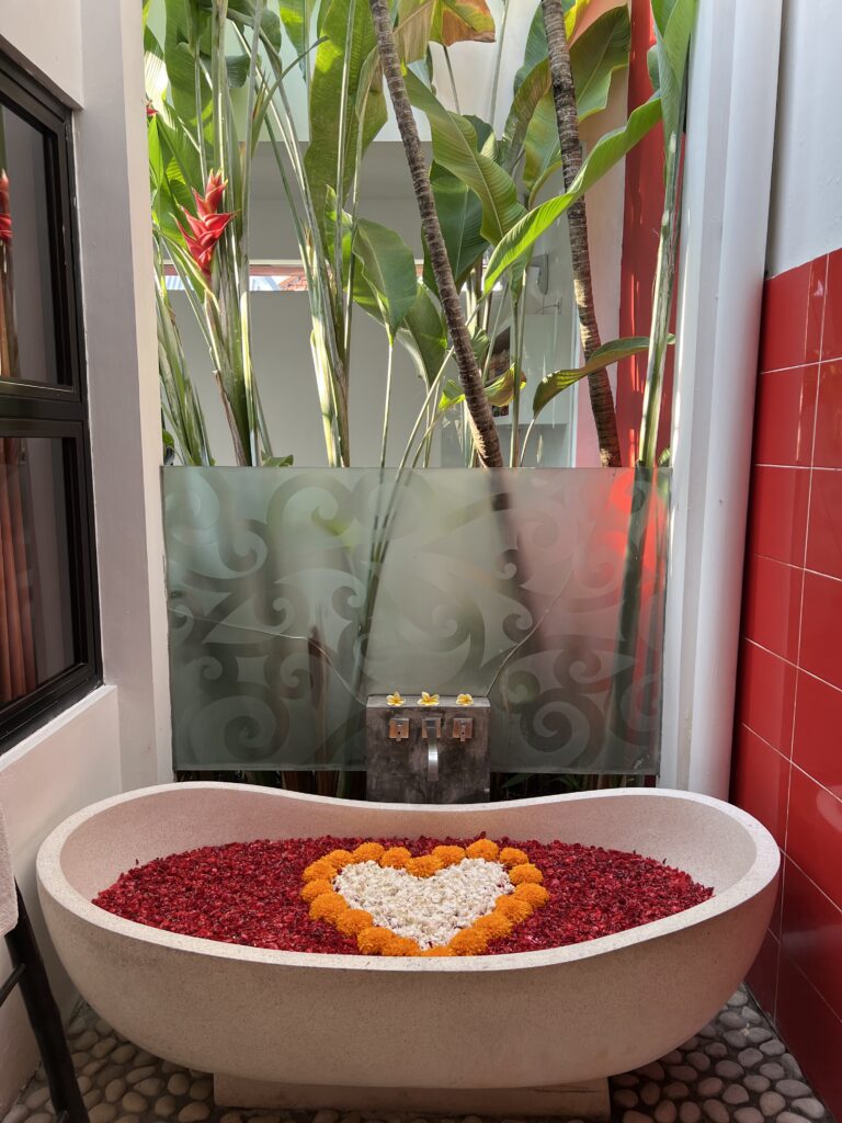 A large tub filled with Bali flowers in the shape of a heart. Background are tropical trees