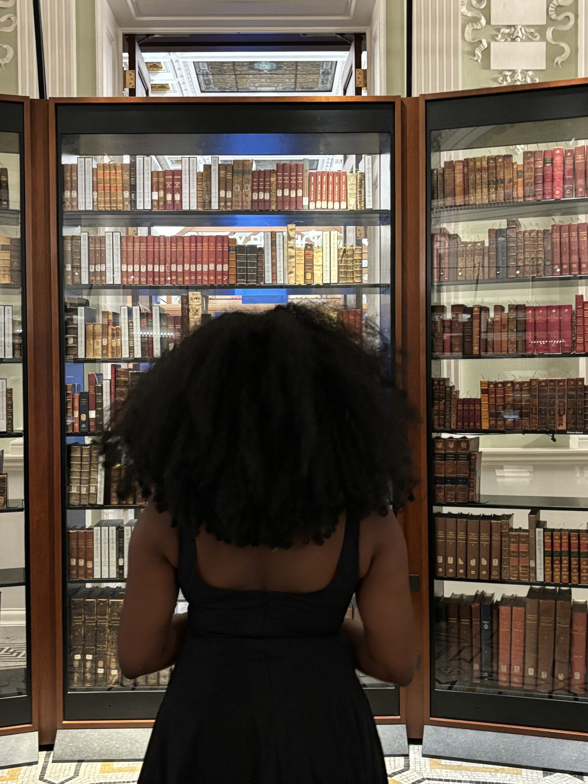 a dark skinned Black woman standing with her back to us, facing a shelf of books