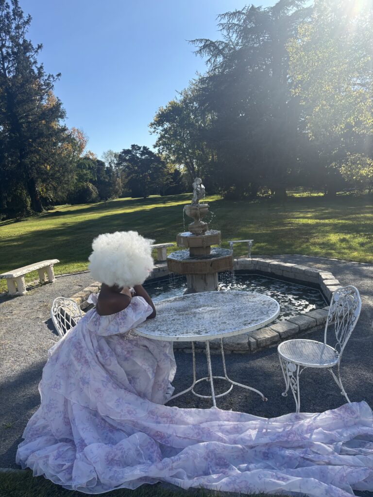 Black woman in long selkie dress sitting at a white table in front of a water fountain at Rosemont Manor