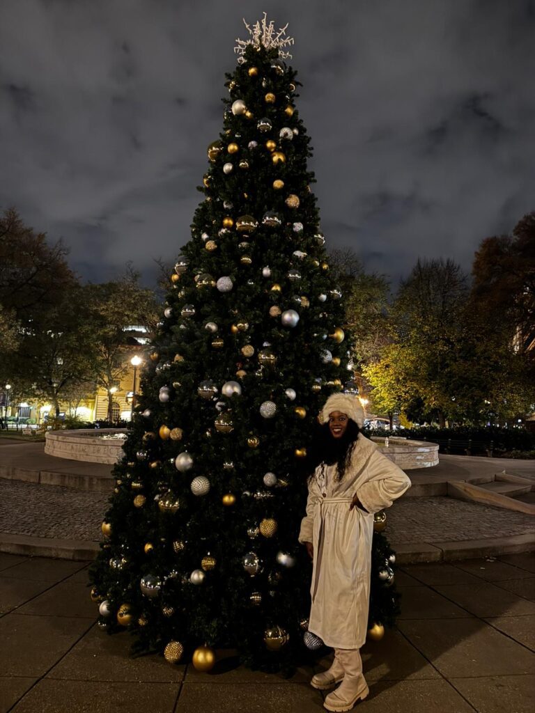 Black woman in white coat and puffy hat standing in front of a lit Christmas Tree in Dupont Circle in Washington DC near the DC Christmas Market