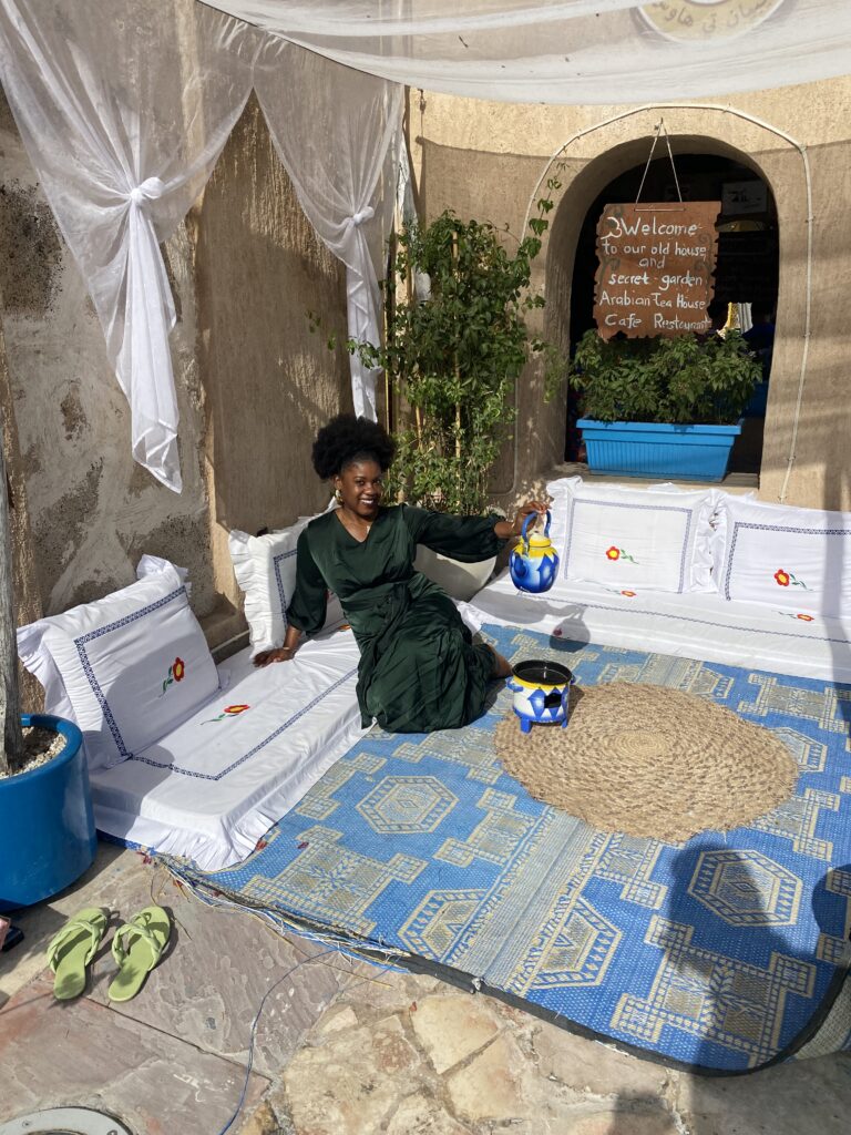 a Black woman with a afro puff wearing a modest green dress while sitting on traditional Emirati chairs on the floor