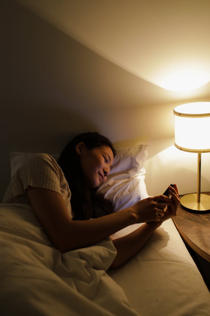 An adult woman casually lying in bed, browsing her smartphone by the dim bedside lamp's light.