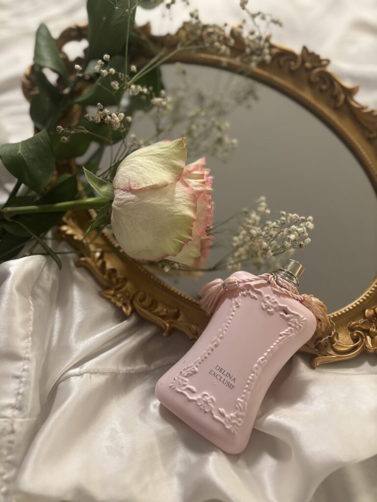 Delina, a feminine pink fragrance bottle, lying on a vintage mirror with flowers surrounding it