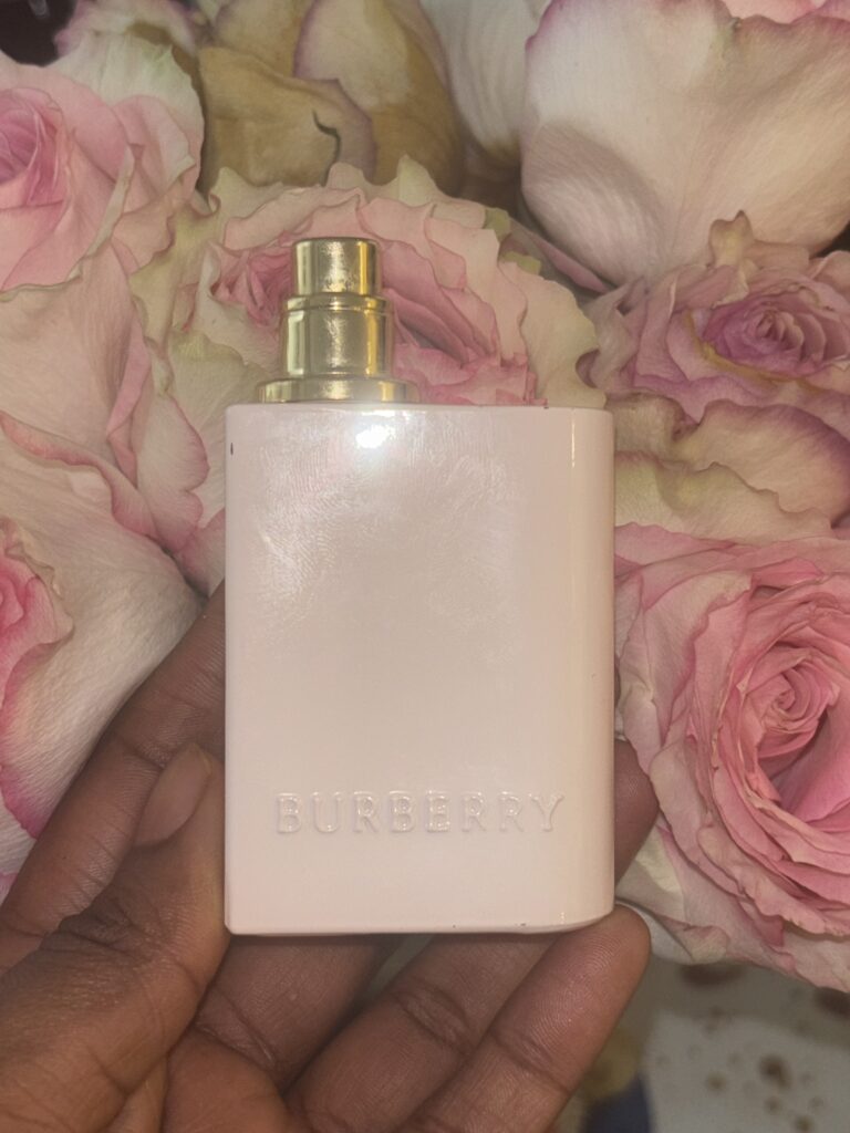 a dark skinned hand holding the Burberry Her perfume in front of pink roses