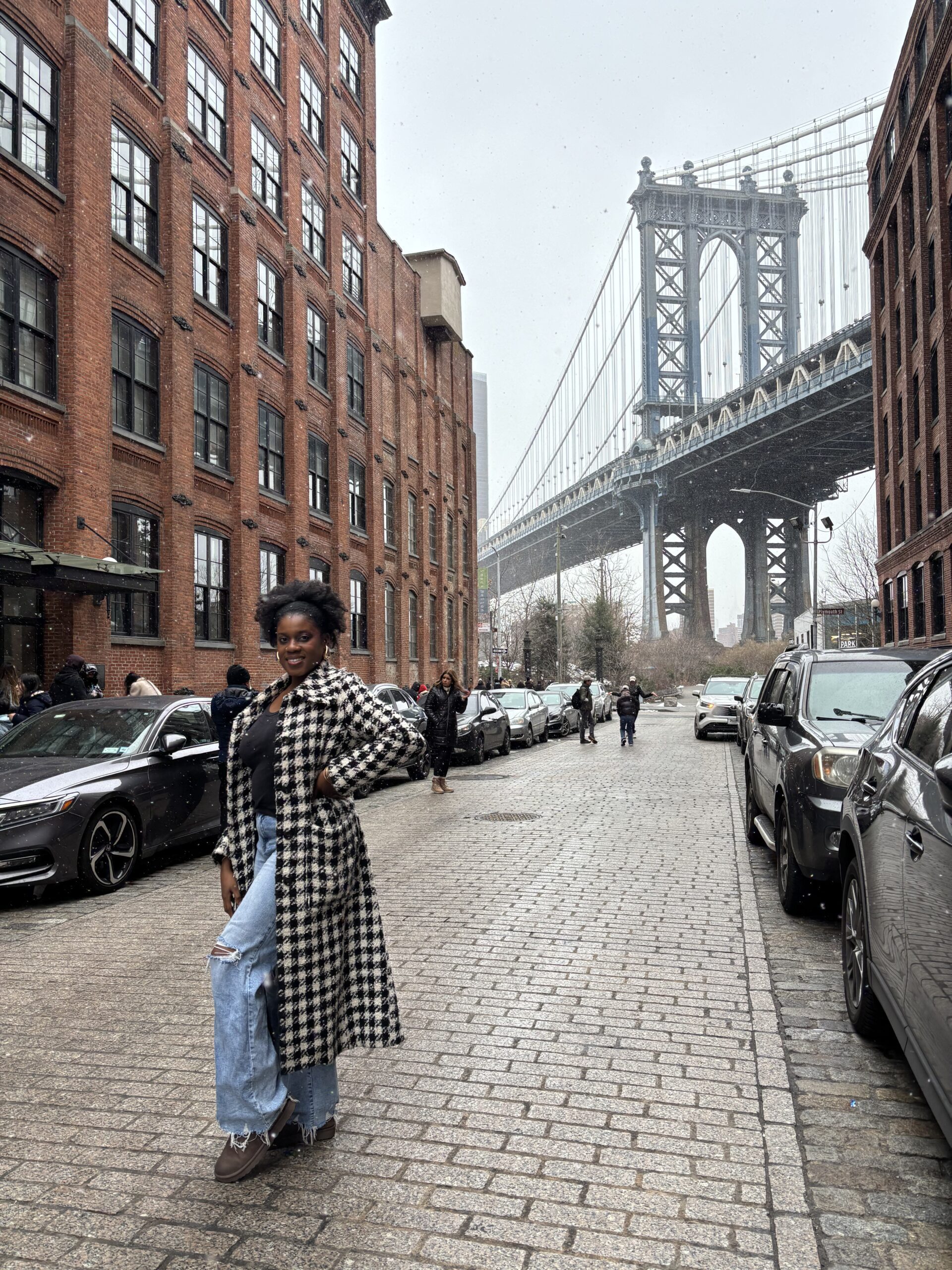 a Black woman in Black and white trench coat standing in front of the DUMBO Manhattan Bridge view