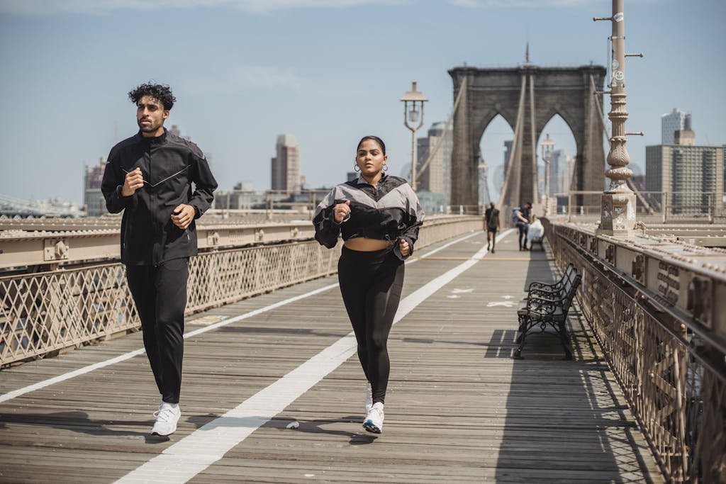 Two adults jogging on Brooklyn Bridge with city skyline in the background, showcasing health and vitality.